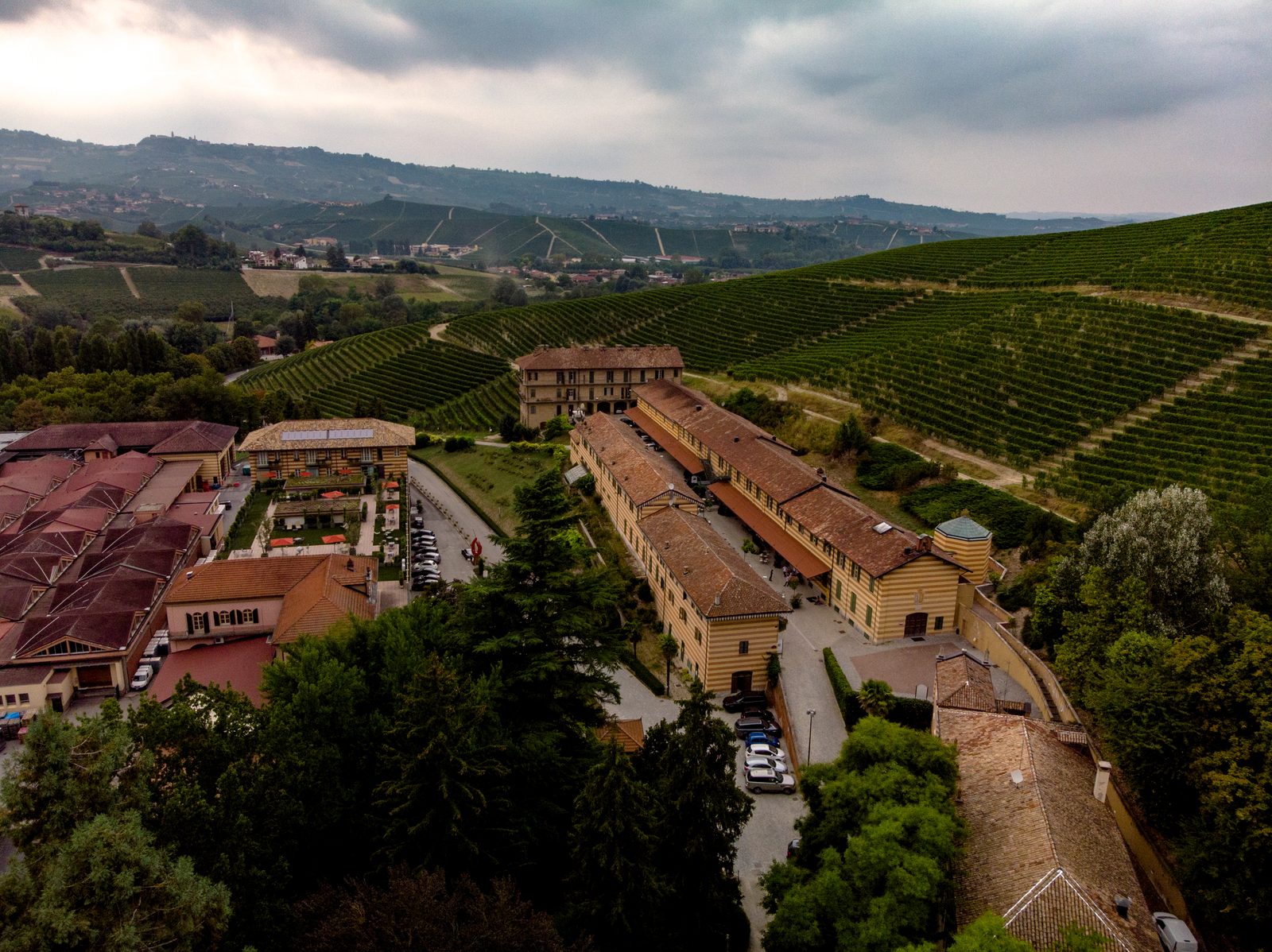 Una tenuta vista dal drone: edifici a strisce rosse e gialle intorno a un cortile, dietro le colline coperte di vigne sotto un cielo nuvoloso.