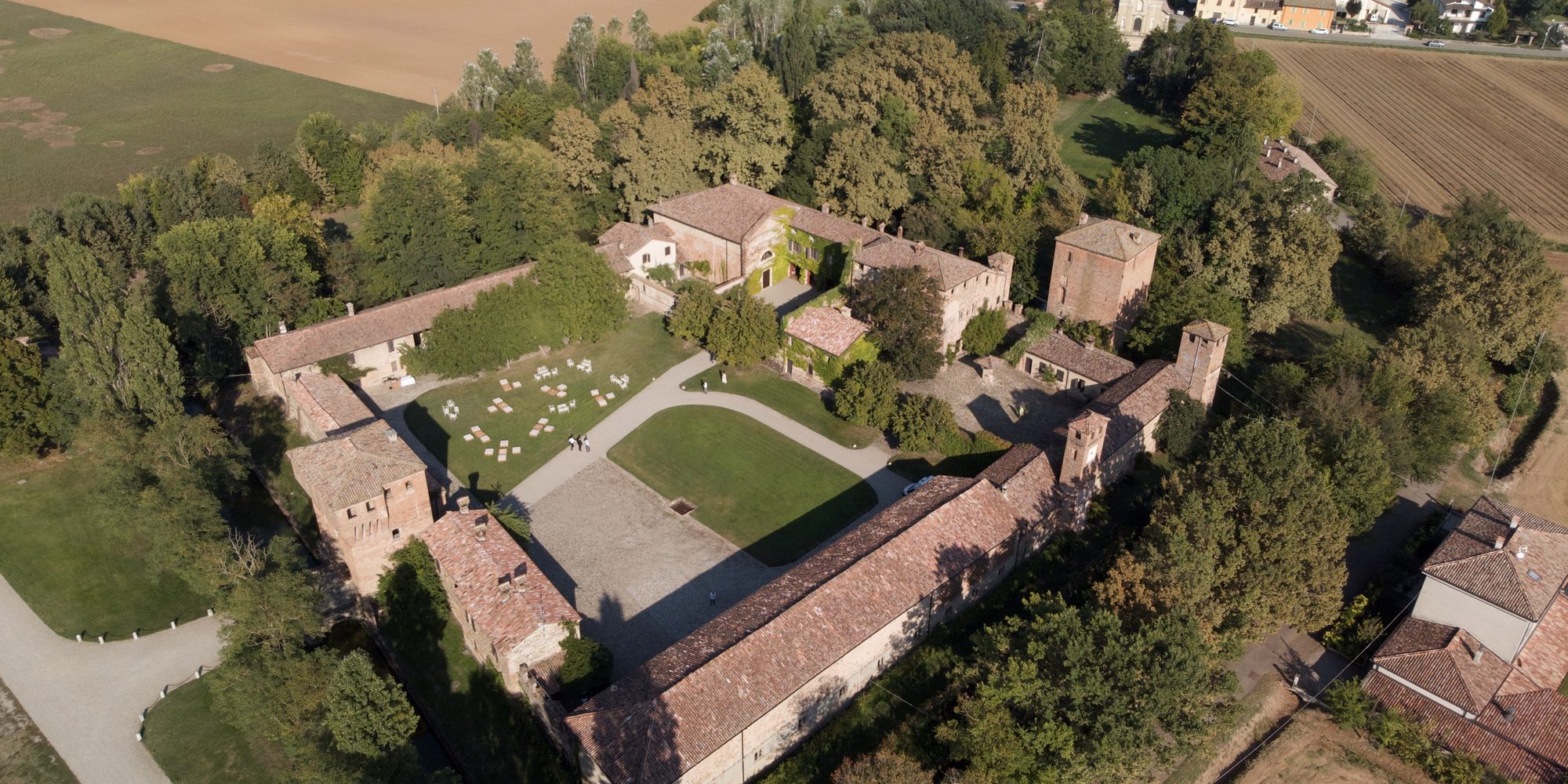 Il Castello di Paderna visto dal drone: le torri, i tetti, il cortile e il prato con le sedie bianche pronte per la cerimonia, la campagna intorno.