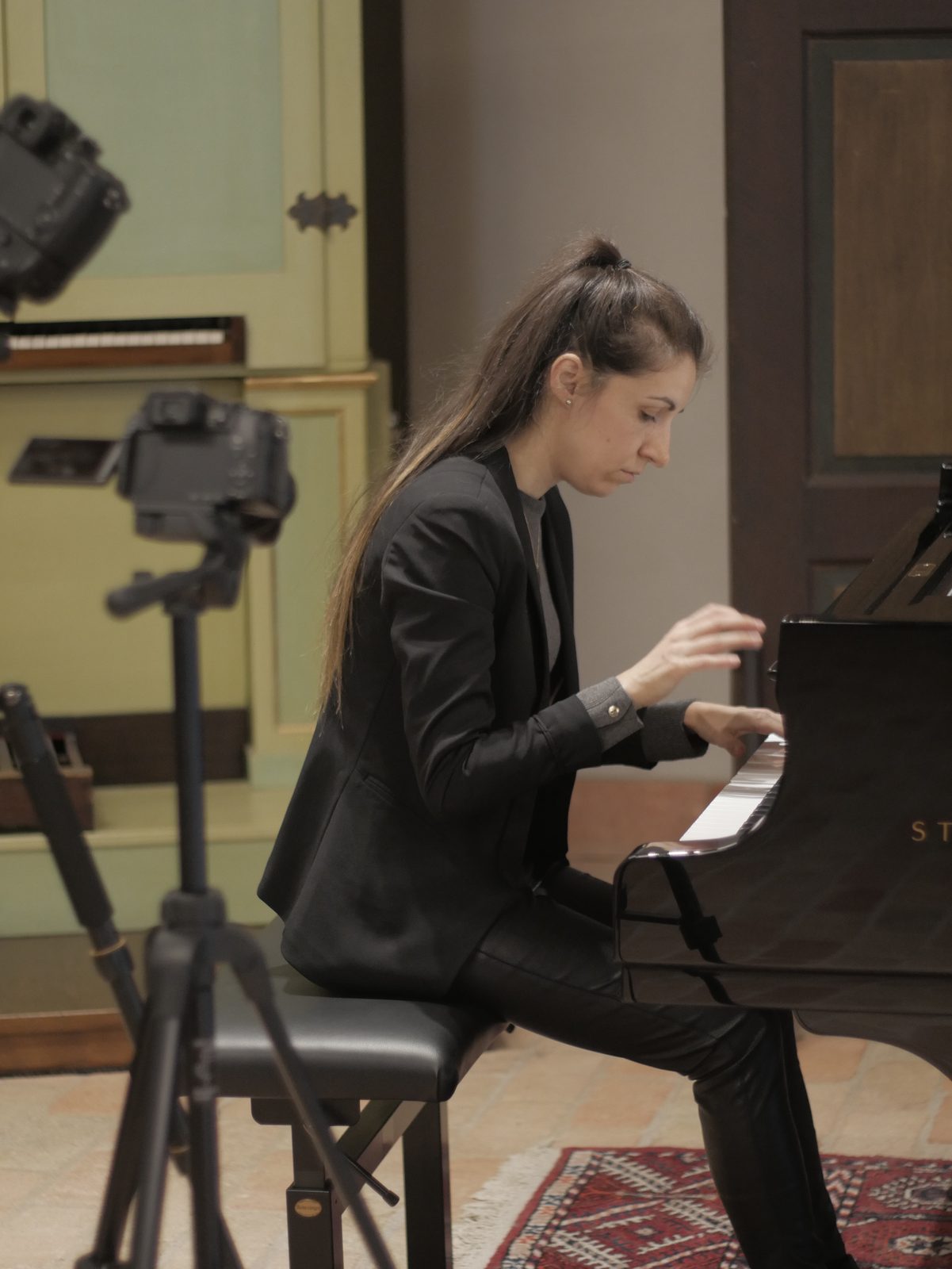 La pianista suona, vista di profilo, su un pianoforte a coda; davanti a lei due macchine fotografiche su treppiede la riprendono. Sullo sfondo un antico strumento a tastiera, con la cassa dipinta di verde chiaro.