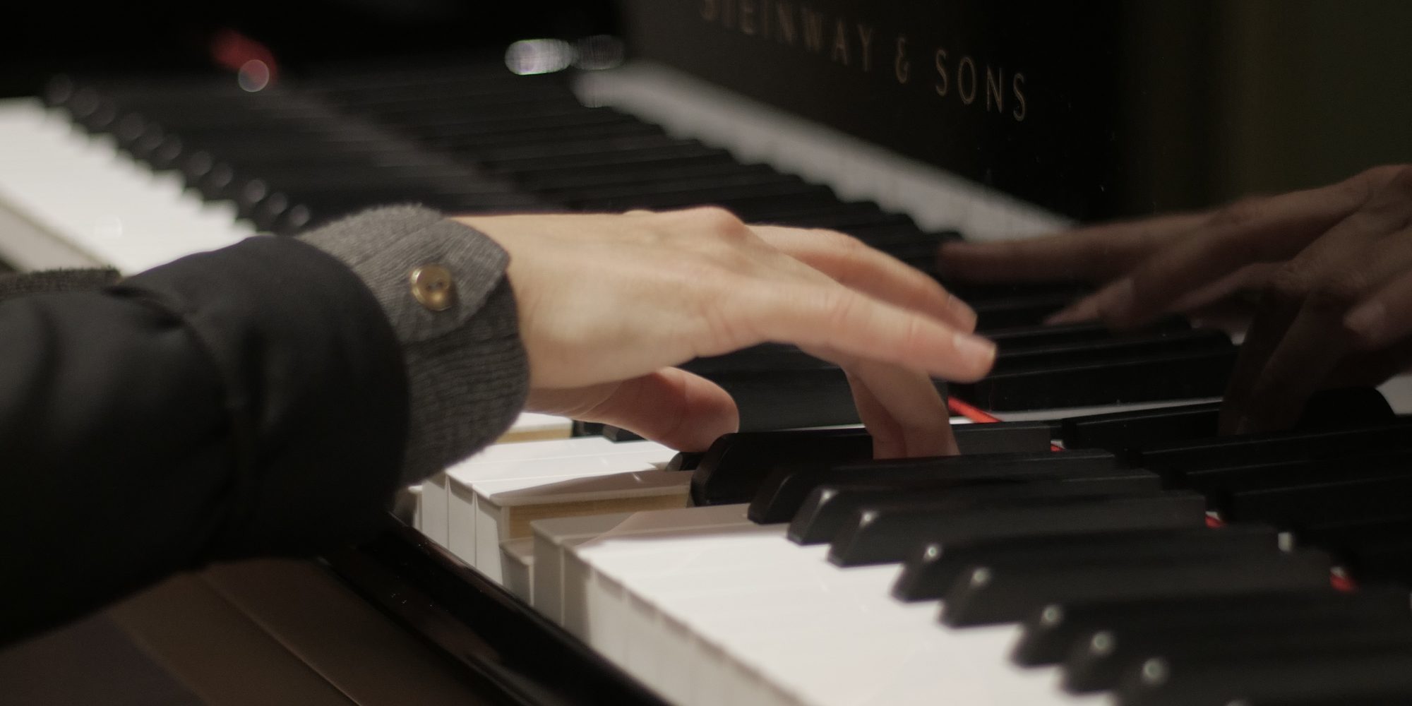 Le mani della pianista sulla tastiera di uno Steinway, viste da vicino, con il riflesso delle dita nel legno lucido sopra i tasti.