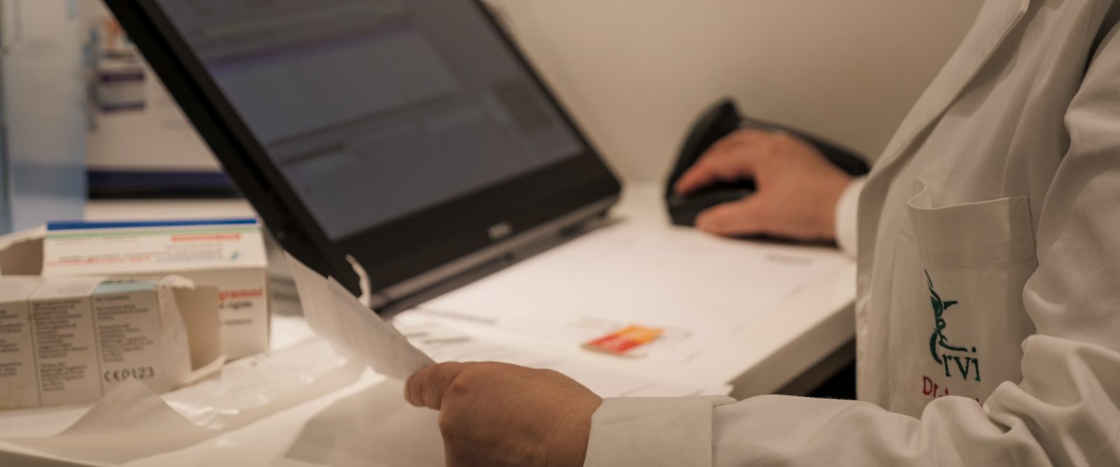 Un banner orizzontale: la manica del camice con il marchio della farmacia, una mano sul mouse e una che tiene un foglio davanti allo schermo, le scatole dei medicinali sfocate in primo piano.