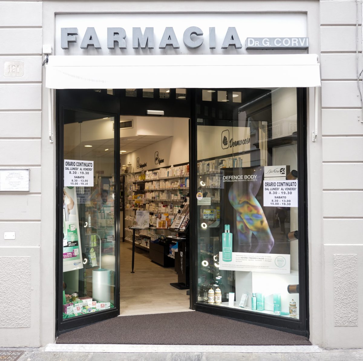 L’ingresso della Farmacia Dr. G. Corvi: l’insegna in lettere d’acciaio, la tenda bianca, le porte a vetri aperte sugli scaffali e i cartelli dell’orario continuato.
