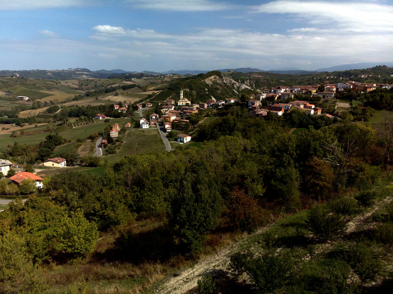 Le colline del Tortonese dal drone: un paese sul crinale, i campi e i boschi, il cielo con nuvole leggere.