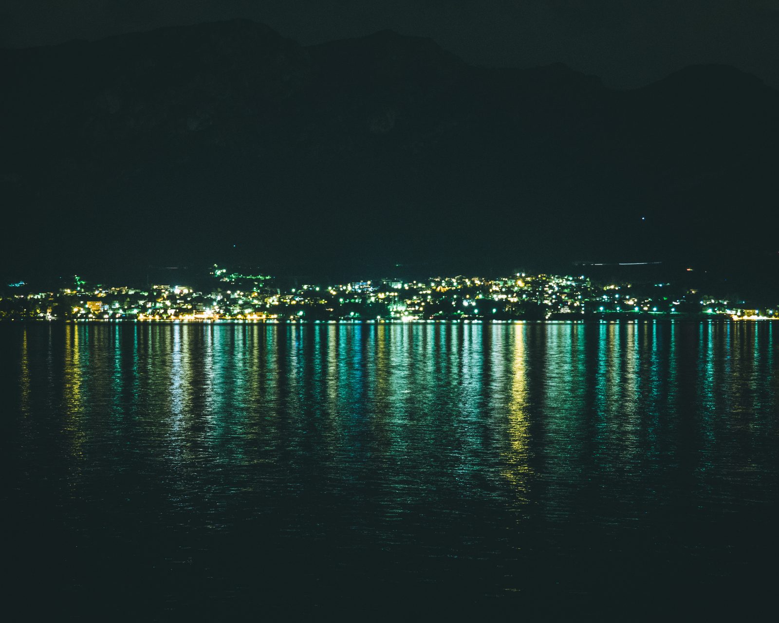 Il lago di notte: le luci di un paese sulla riva opposta si riflettono nell’acqua in lunghe strisce verdi e gialle, sotto le montagne scure.