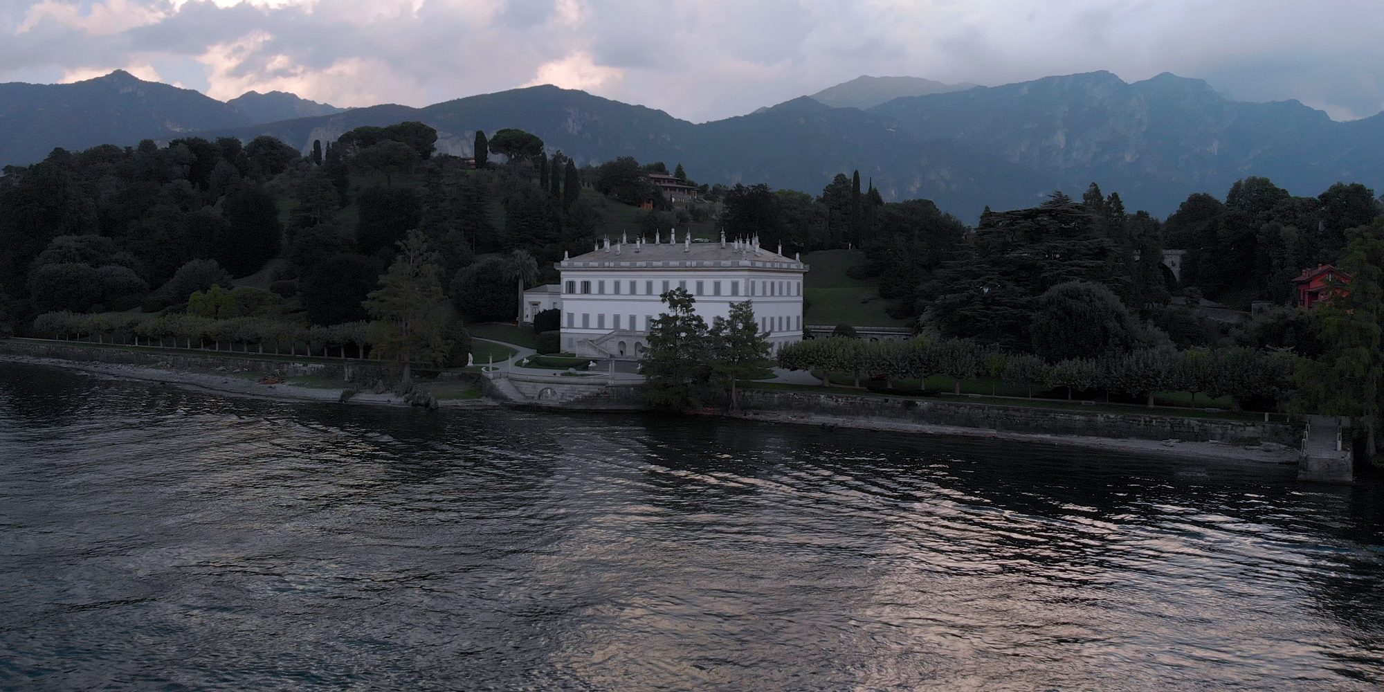 Una villa bianca sulla riva del lago, ripresa dal drone sull’acqua la sera; dietro, il parco con i pini e le montagne sotto le nuvole.