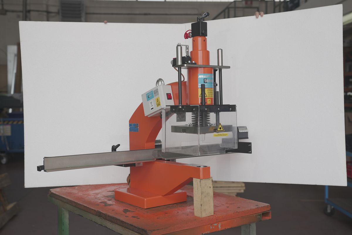 An orange hydraulic drilling machine on a workshop bench; behind it, a large white panel held up by hand; around it, the shop floor with shelves and trolleys.