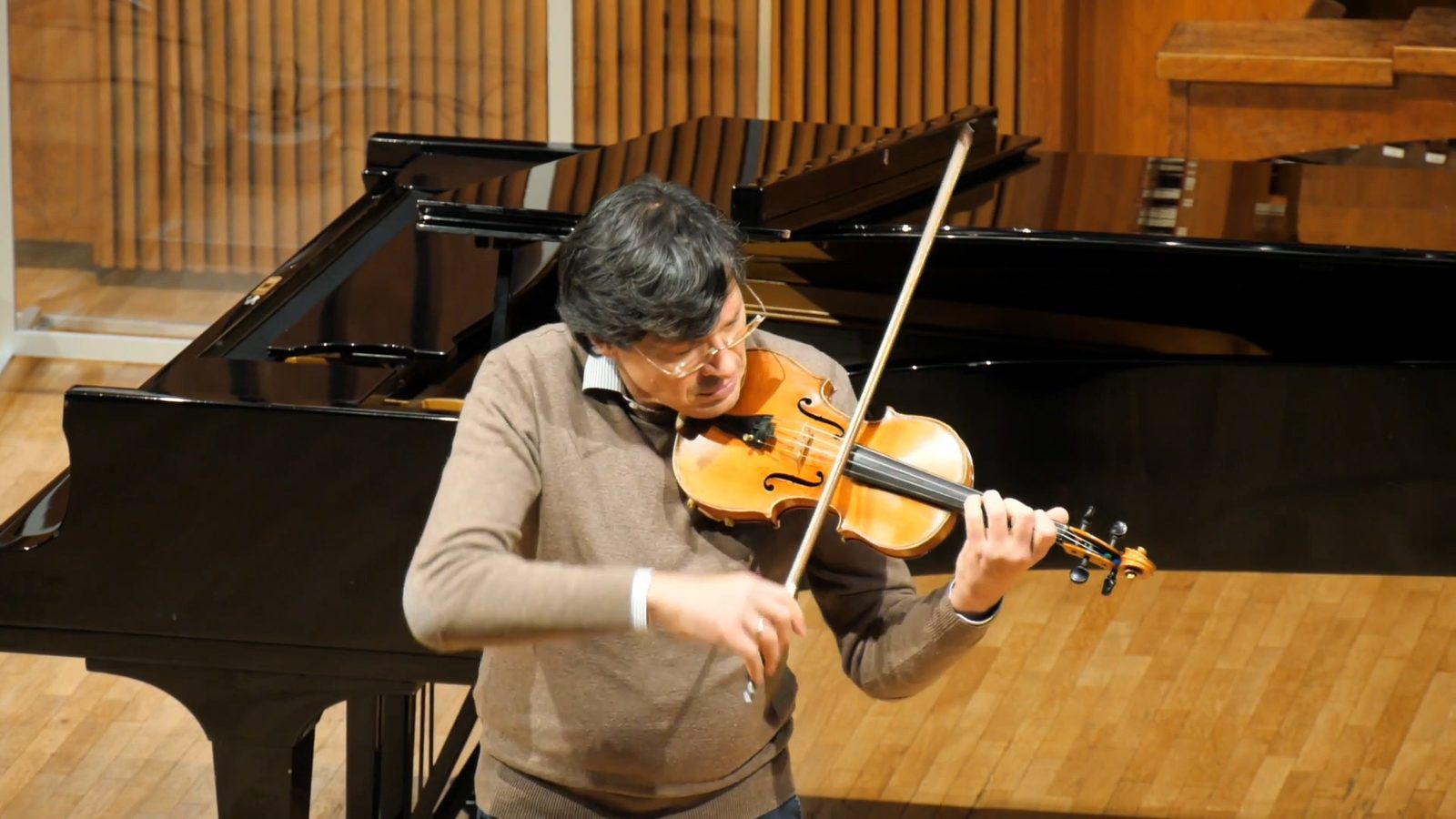 Un violinista in maglione suona in piedi davanti a un pianoforte a coda, sul palco di legno, con l’arco alzato.