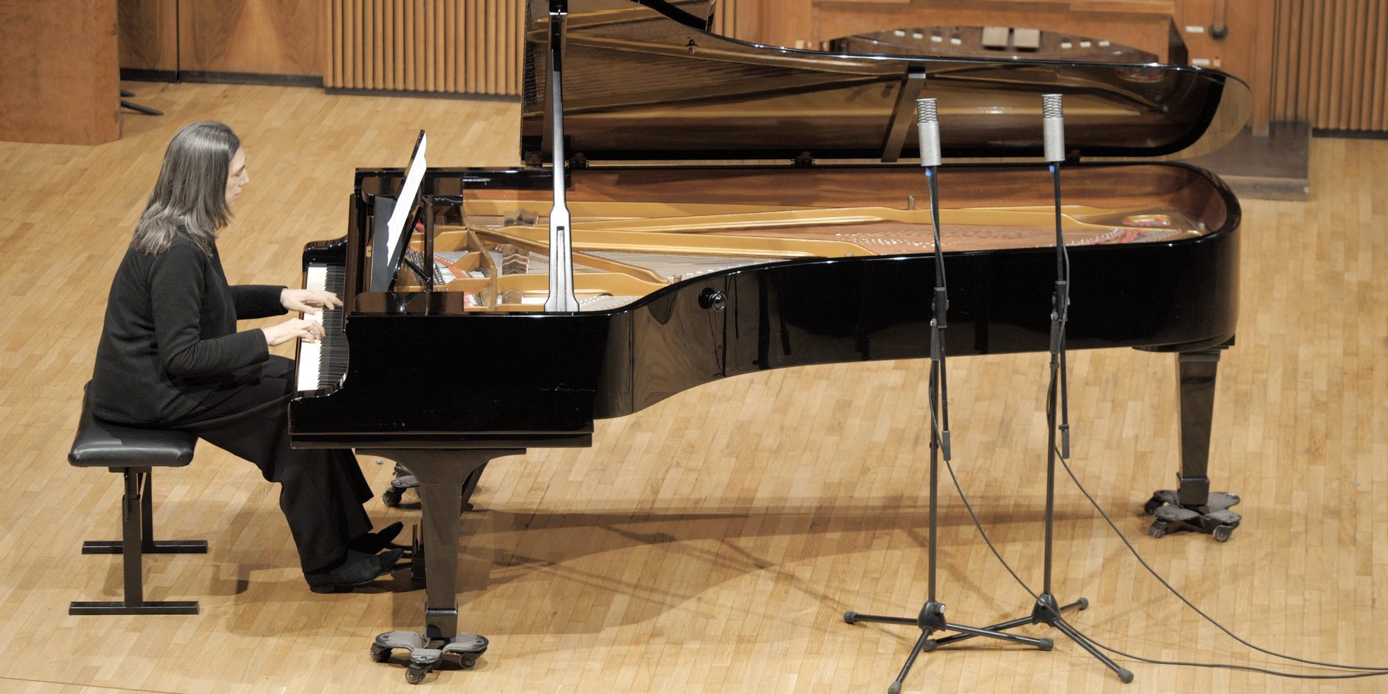 Una pianista suona un pianoforte a coda aperto sul palco di legno del Salone del Nicolini, ripresa dall’alto; davanti alla coda due microfoni su aste, dietro le doghe di legno e la consolle dell’organo.