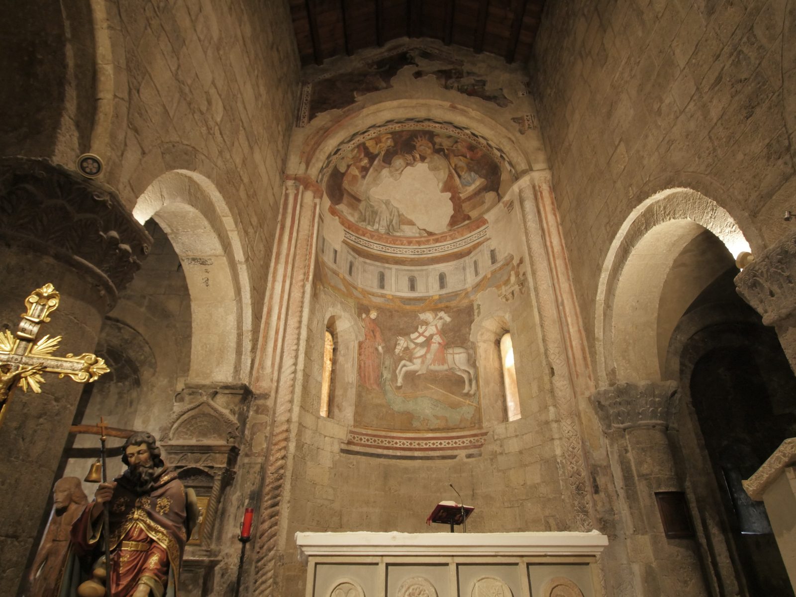 The frescoed apse of the church, with a knight on a white horse in the middle, between two narrow windows; in front, the pale altar, a wooden statue and a gilded cross.