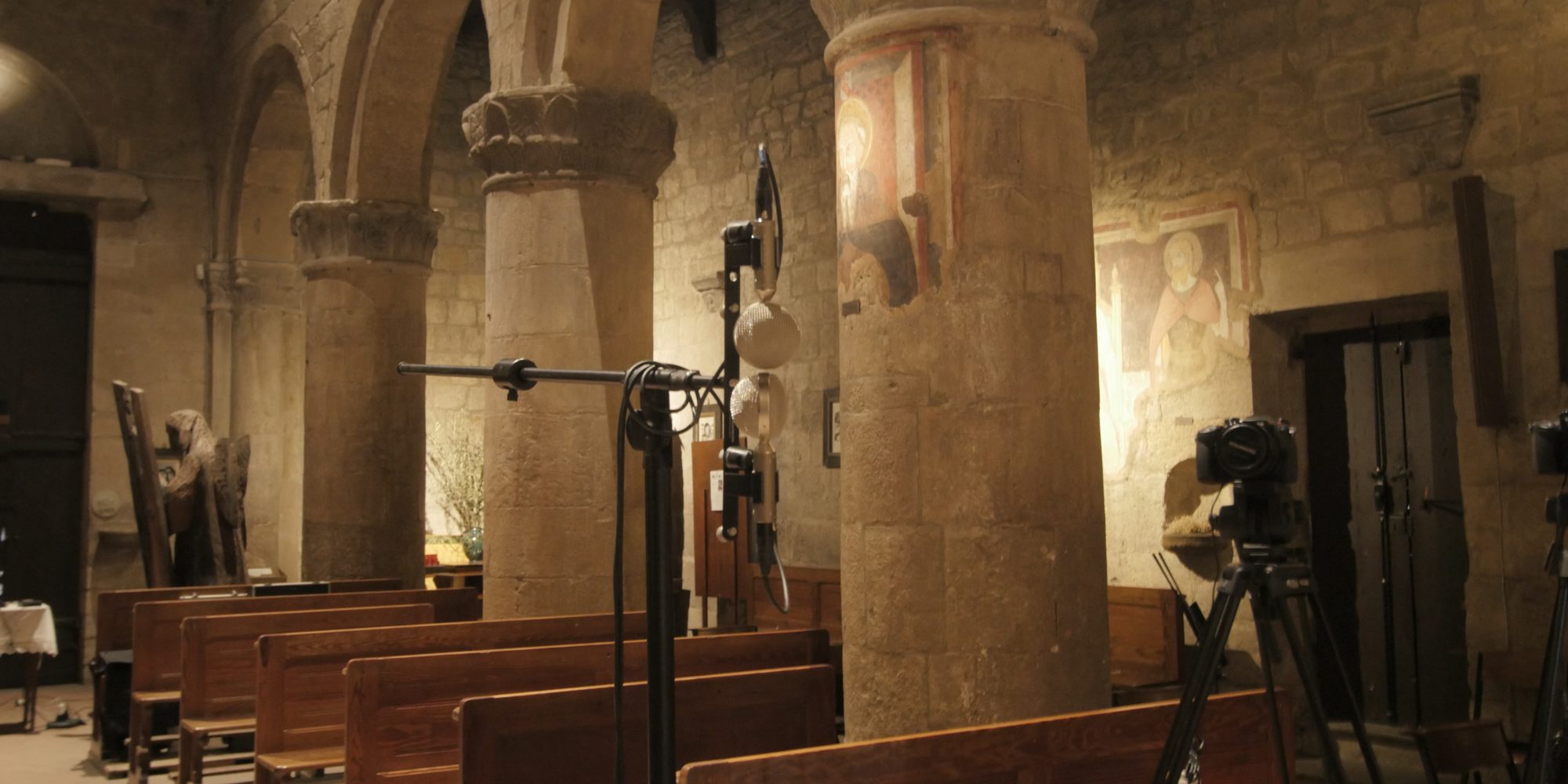 La navata della pieve di Vigoleno con le colonne di pietra e gli affreschi; fra i banchi di legno un’asta con i microfoni e due camere sui treppiedi.