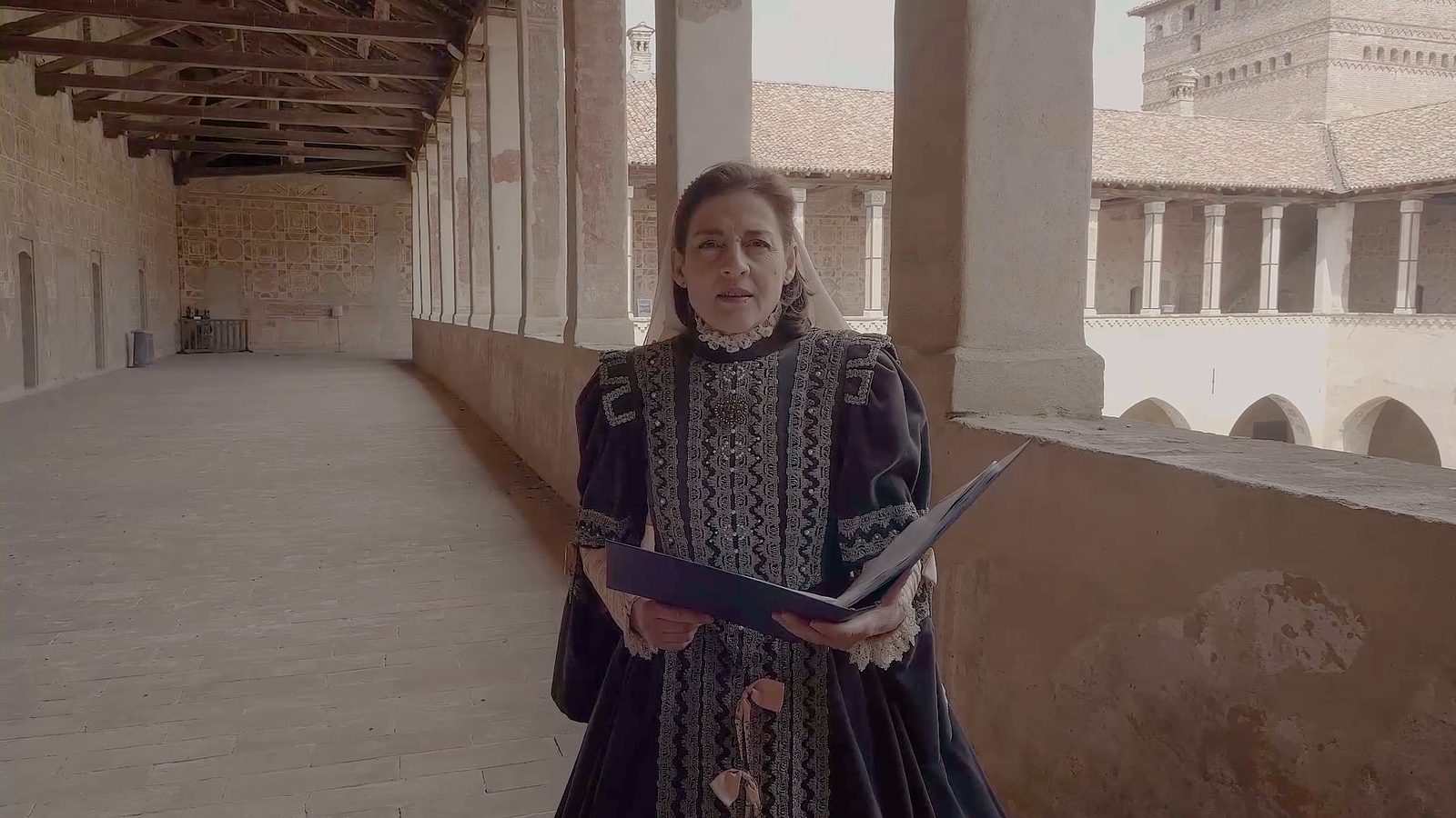 Una cantante in abito scuro ricamato legge da una cartellina nella loggia del castello; alle sue spalle il cortile porticato e la torre di mattoni.