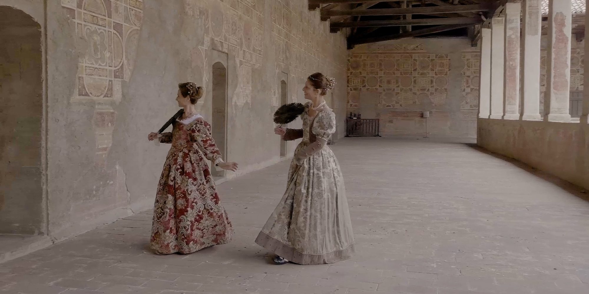 Two dancers in Renaissance gowns, one red and one ivory, with feather fans, turning in the castle loggia between walls painted in panels and the beamed ceiling.