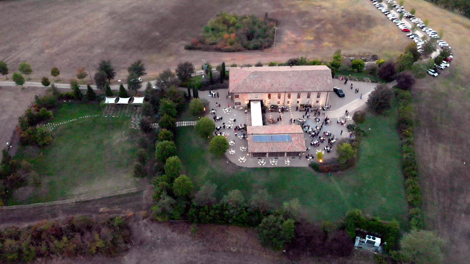 A farmhouse seen from the drone at dusk: the roofs, the courtyard with the tables and the lights on, the garden closed by a hedge, ploughed fields around.