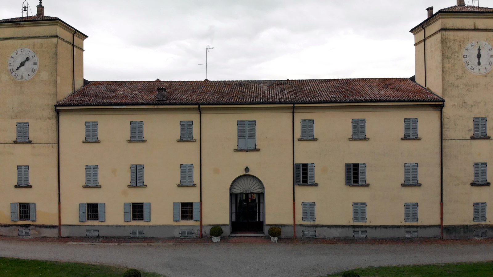 The yellow façade of a villa with grey shutters and an arched doorway, between two clock towers, under an overcast sky.