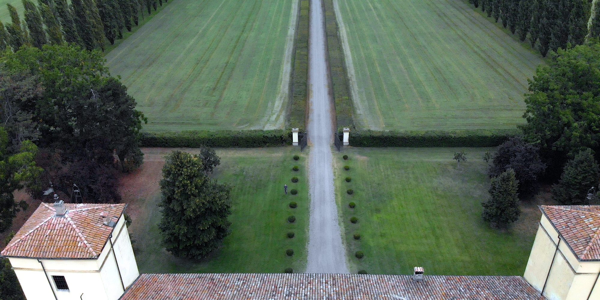 A long white avenue between two mown lawns and rows of trees, seen from the drone above the roof of a villa, between its two towers.