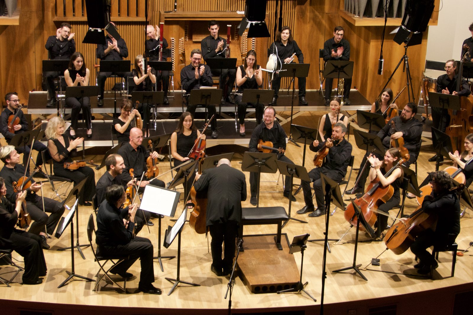 L’orchestra a fine concerto, vista dall’alto della sala: i musicisti applaudono in piedi e seduti, il direttore di spalle sul podio, i leggii e i microfoni sulle aste.