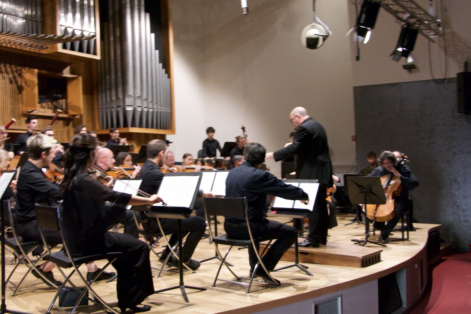 La prova del pomeriggio nel Salone: il direttore in piedi davanti all’orchestra, gli archi in primo piano, l’organo sul fondo, l’americana con i fari in alto.