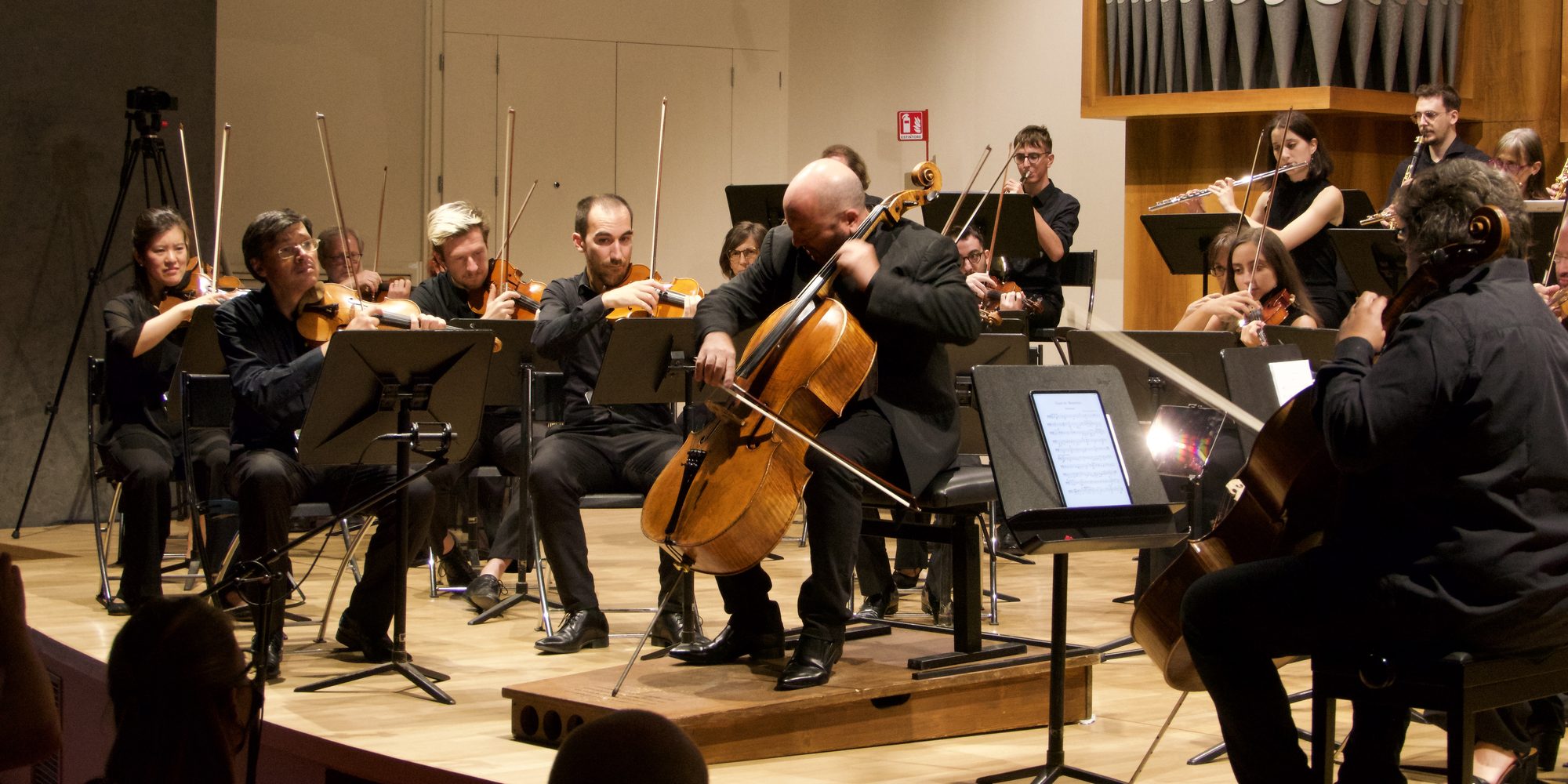 Enrico Dindo suona il violoncello al centro del palco del Salone del Nicolini, in mezzo all’orchestra che accompagna; dietro, le canne dell’organo; a sinistra una camera sul treppiede.
