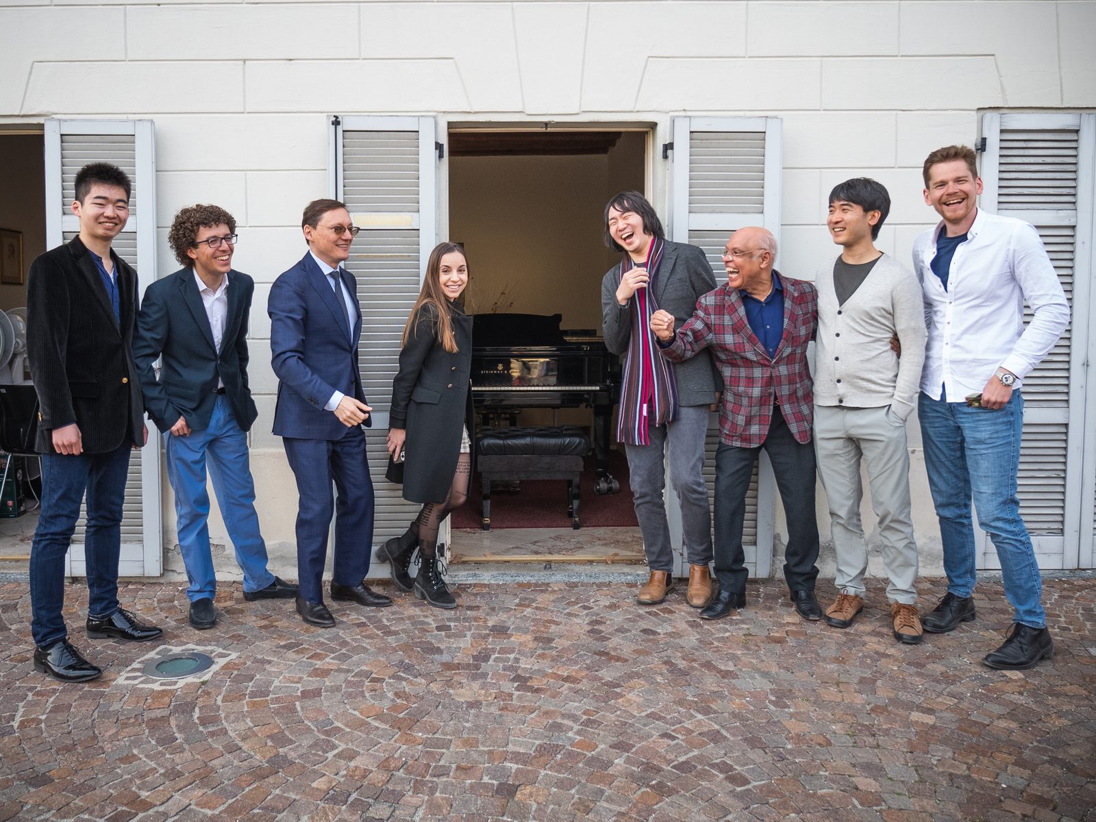 Teachers and students standing in front of the Academy’s villa, on the cobbles, between the white shutters; through the open door you can see the pianos. Some of them are laughing.