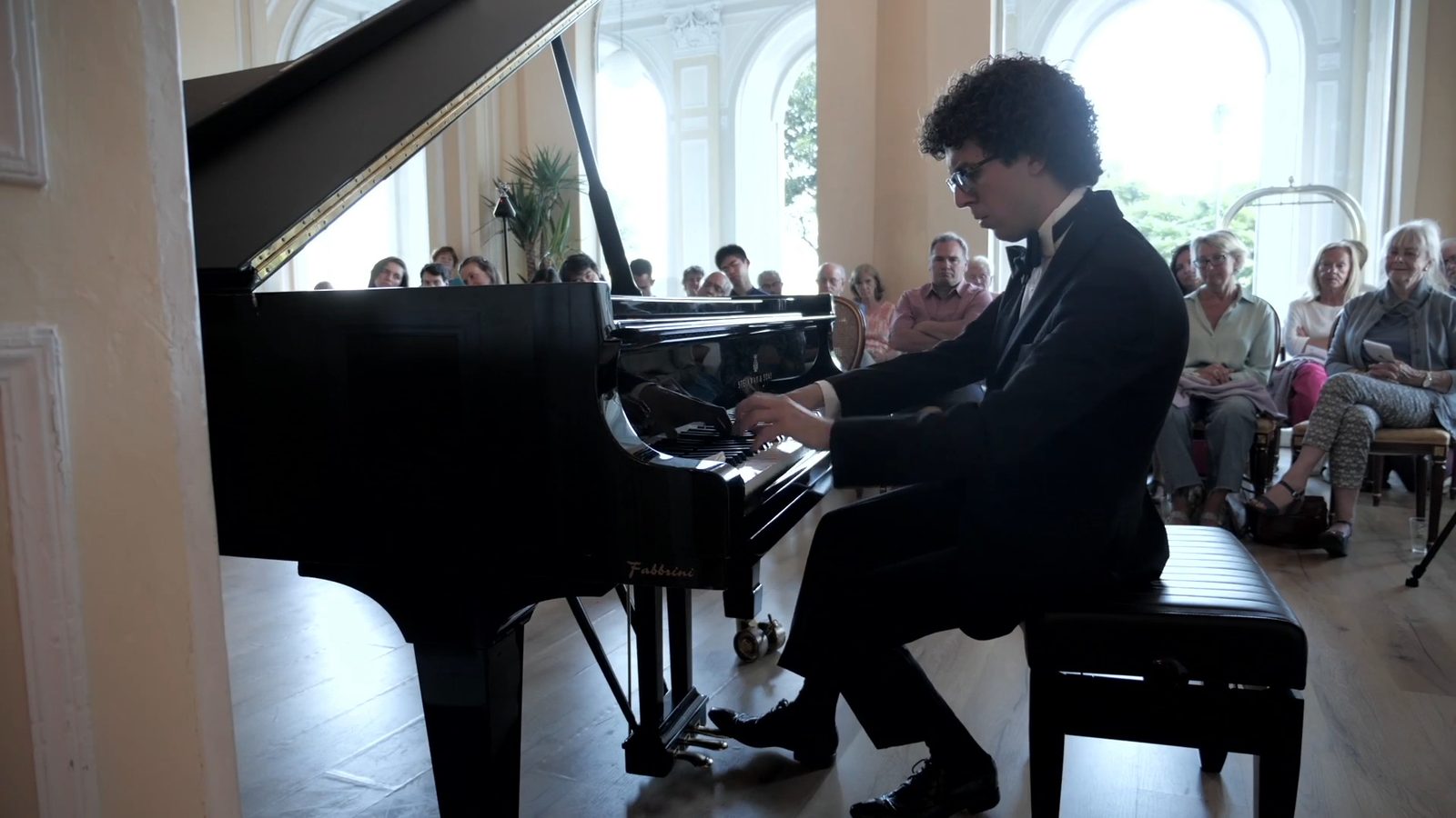 A young pianist in tails plays a grand piano, filmed from the side; behind him the seated audience and the large arched windows with the light of the lake.