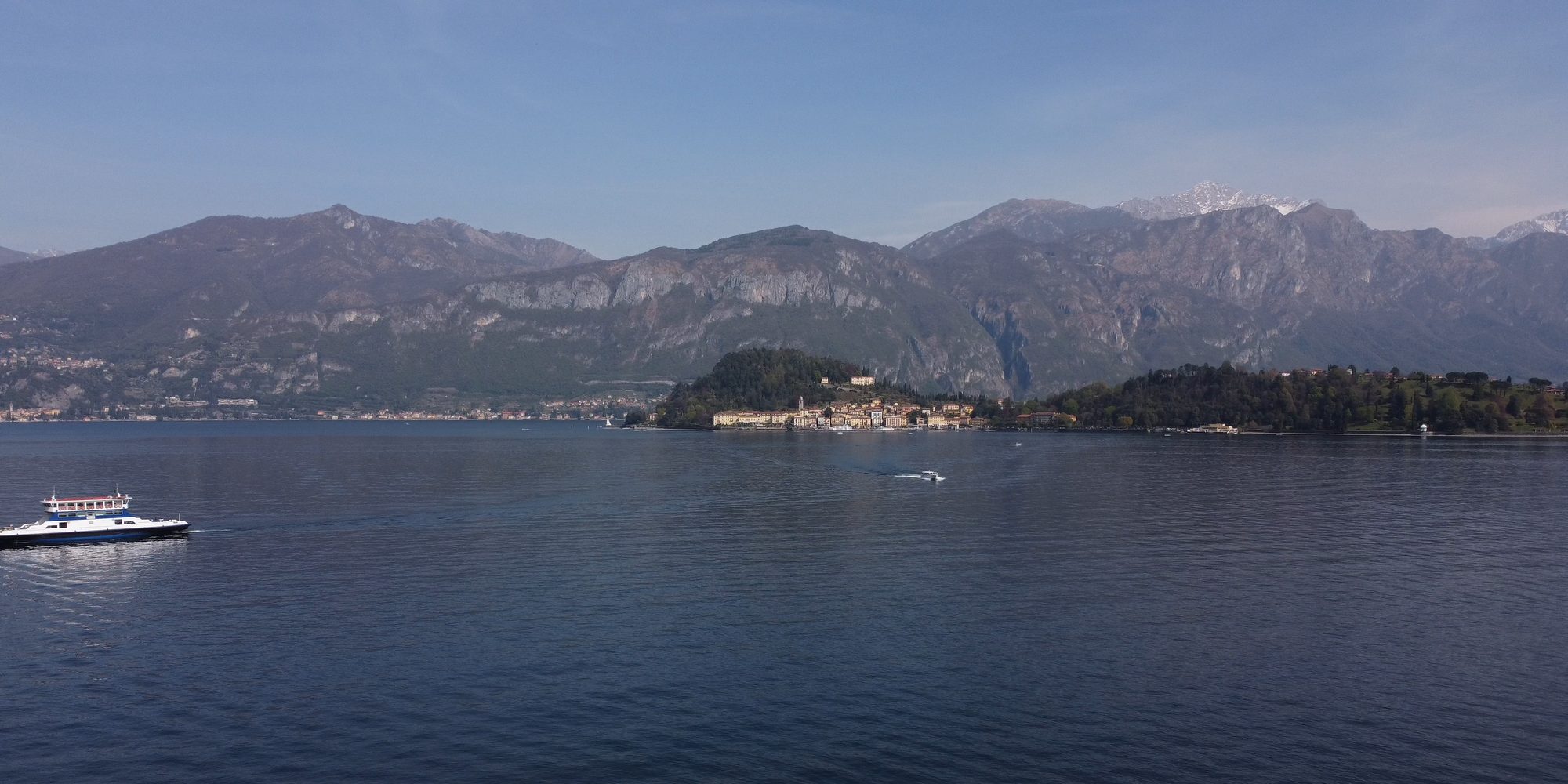 Il Lago di Como ripreso dal drone in primavera: l’acqua blu, un traghetto bianco a sinistra, il promontorio di Bellagio con le case chiare e, dietro, le montagne con le cime innevate.