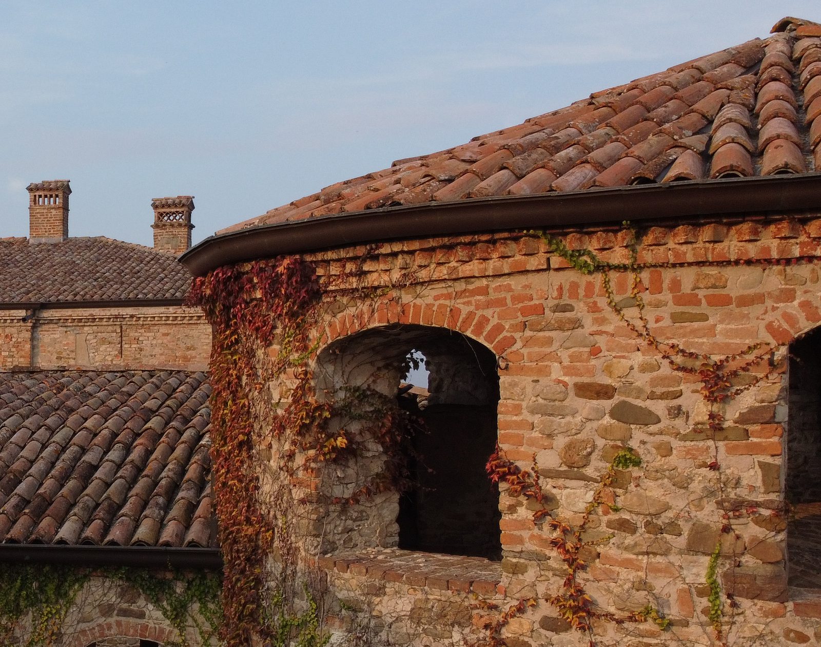 La torre tonda di un castello in mattoni e sassi, coperta dalla vite americana rossa d’autunno, con il tetto di coppi e un arco aperto.