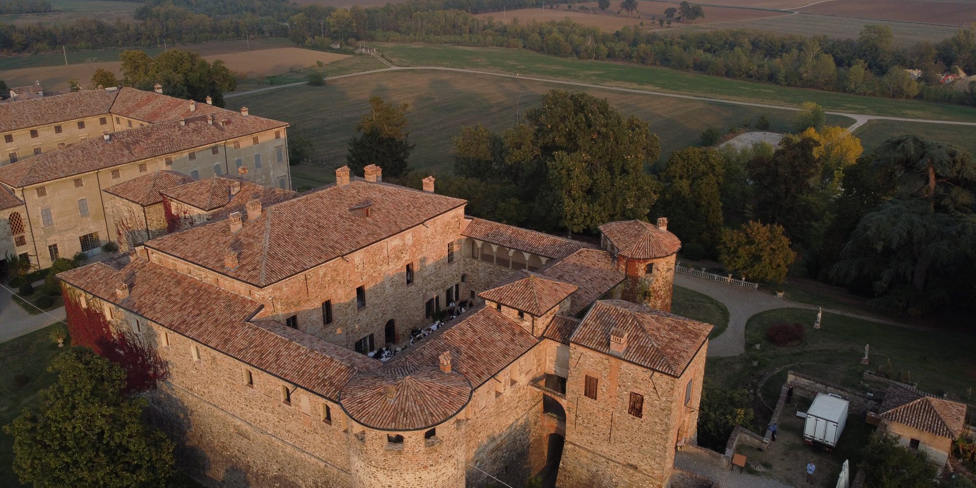 Un castello di mattoni con le torri tonde e il cortile interno, visto dal drone nella luce bassa della sera; intorno gli alberi e la pianura.
