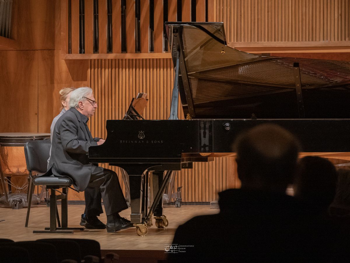 A white-haired pianist at the grand piano, on the wood-panelled stage; in the foreground, backlit, the heads of the audience.