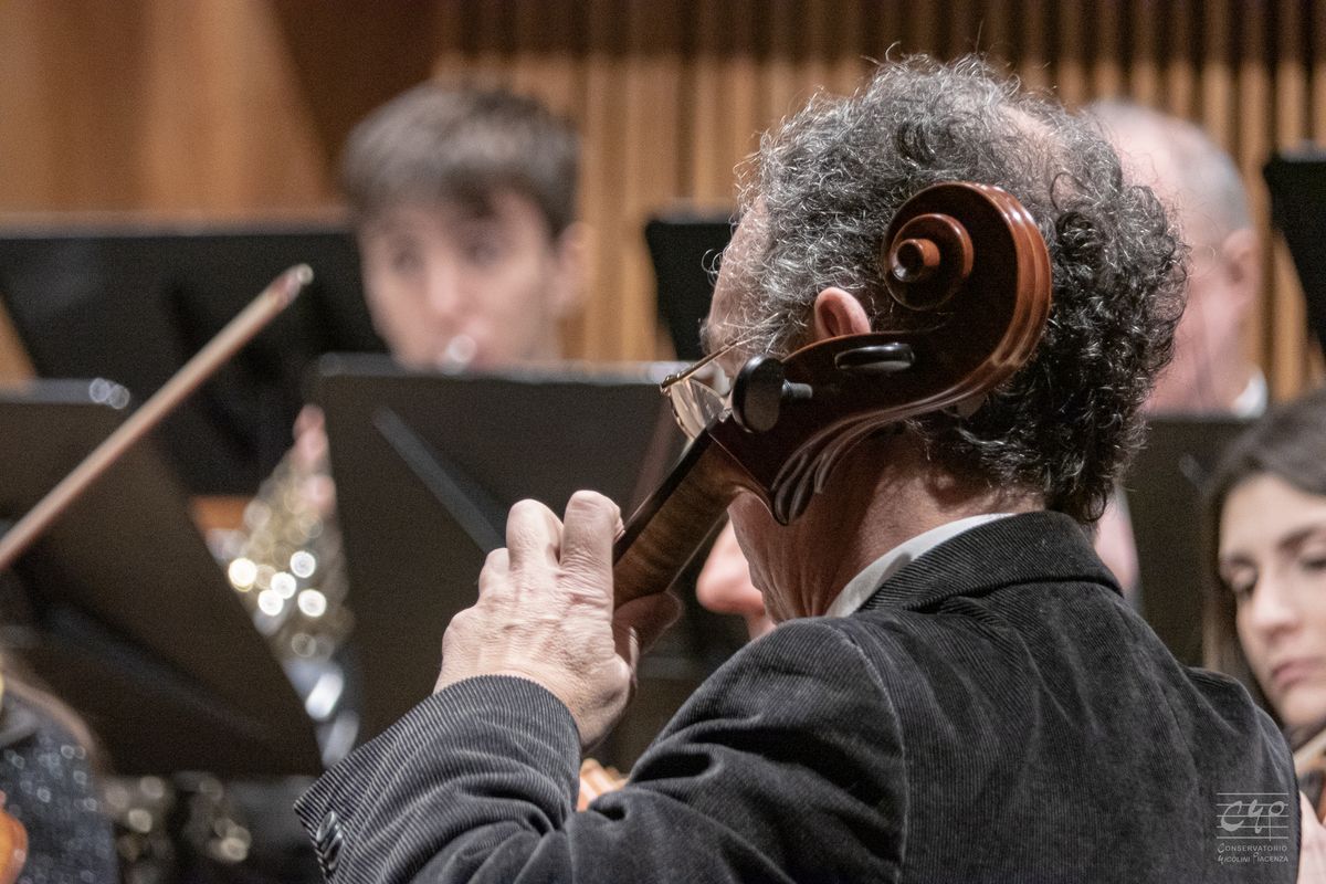 A grey-haired cellist playing in the foreground; behind him the orchestra and the music stands.