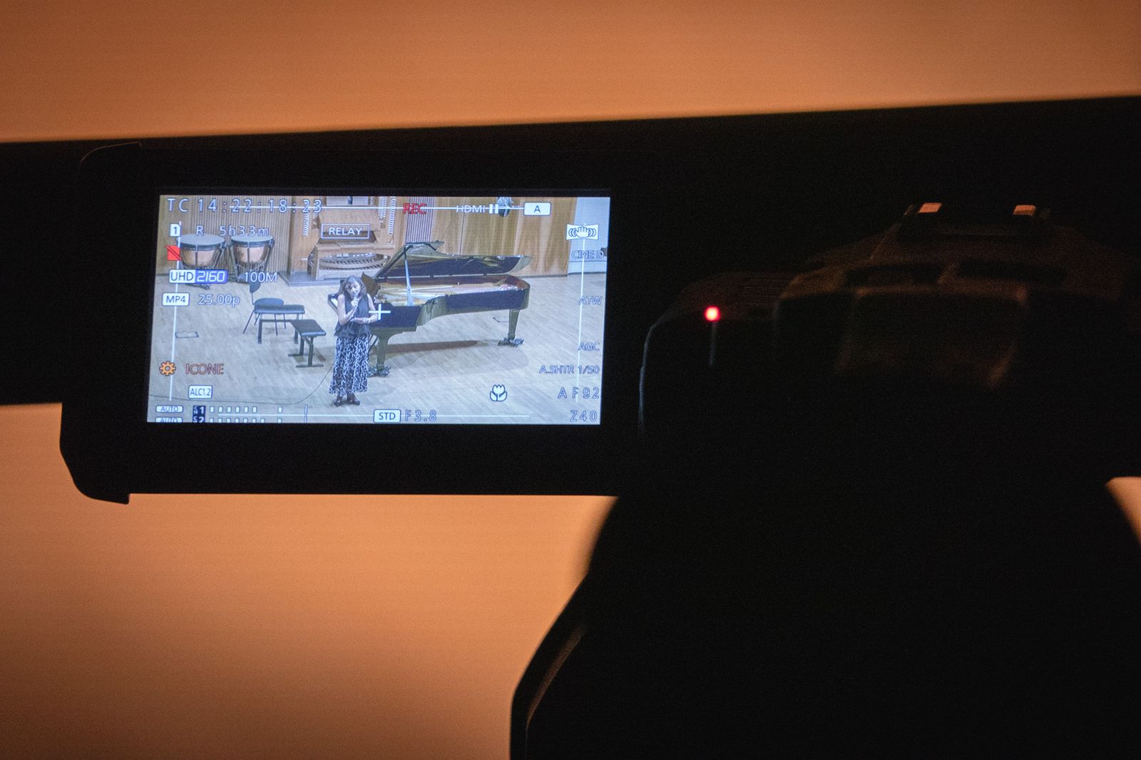 Al buio, in primo piano, il monitor di una videocamera inquadra il palco della sala del Conservatorio con il pianoforte.