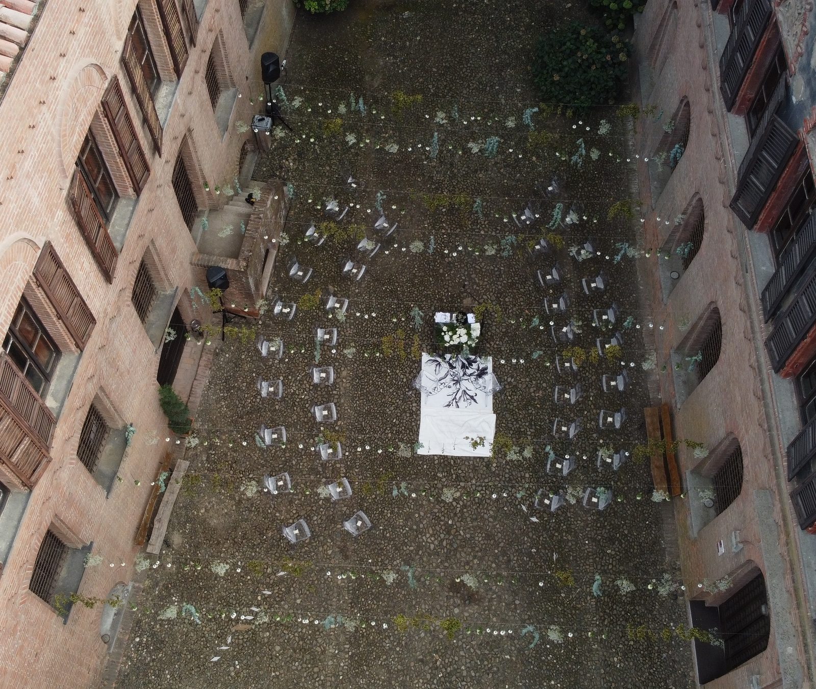 Un cortile acciottolato visto dall’alto, pronto per la cerimonia: le sedie trasparenti in file, i rametti verdi e i fiori bianchi, i fili di luci, il tavolo con il telo bianco al centro.
