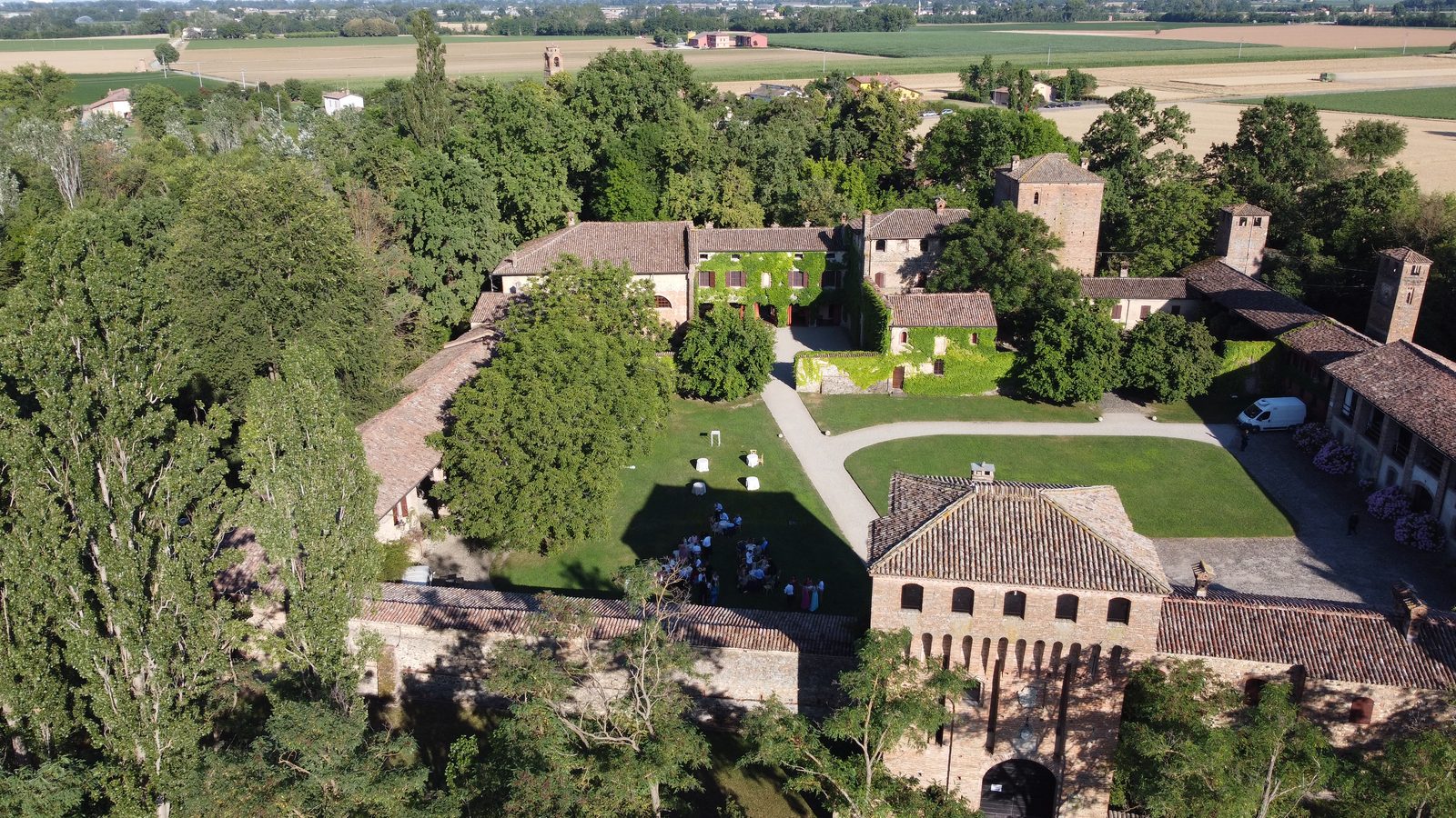 Il Castello di Paderna dal drone in un pomeriggio d’estate: le torri, i tetti, il prato del cortile, gli alberi alti tutto intorno e i campi.