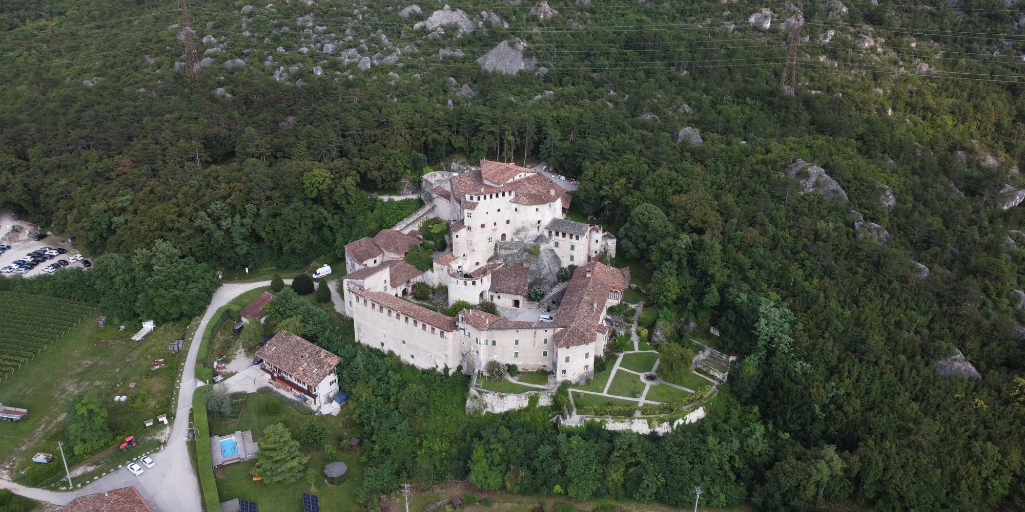 Un castello chiaro con torri e mura sul fianco boscoso di una valle, visto dal drone; sotto, la strada, le vigne e un parcheggio.