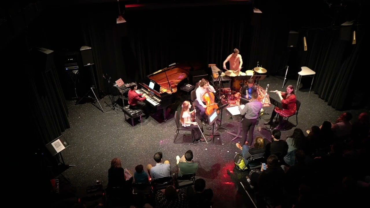 La Dancz Hall dall’alto durante un concerto: un pianoforte, un violoncello, percussioni e altri strumenti al centro della sala nera, il pubblico intorno.