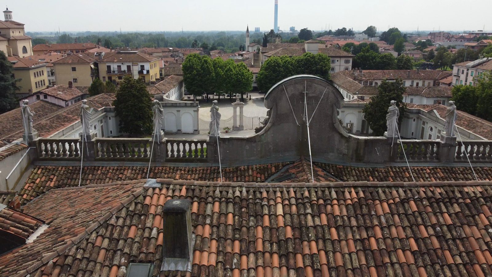 I tetti di coppi della villa visti dal drone, con la balaustra e le statue sul cornicione; intorno le case del paese e, lontano, la pianura.