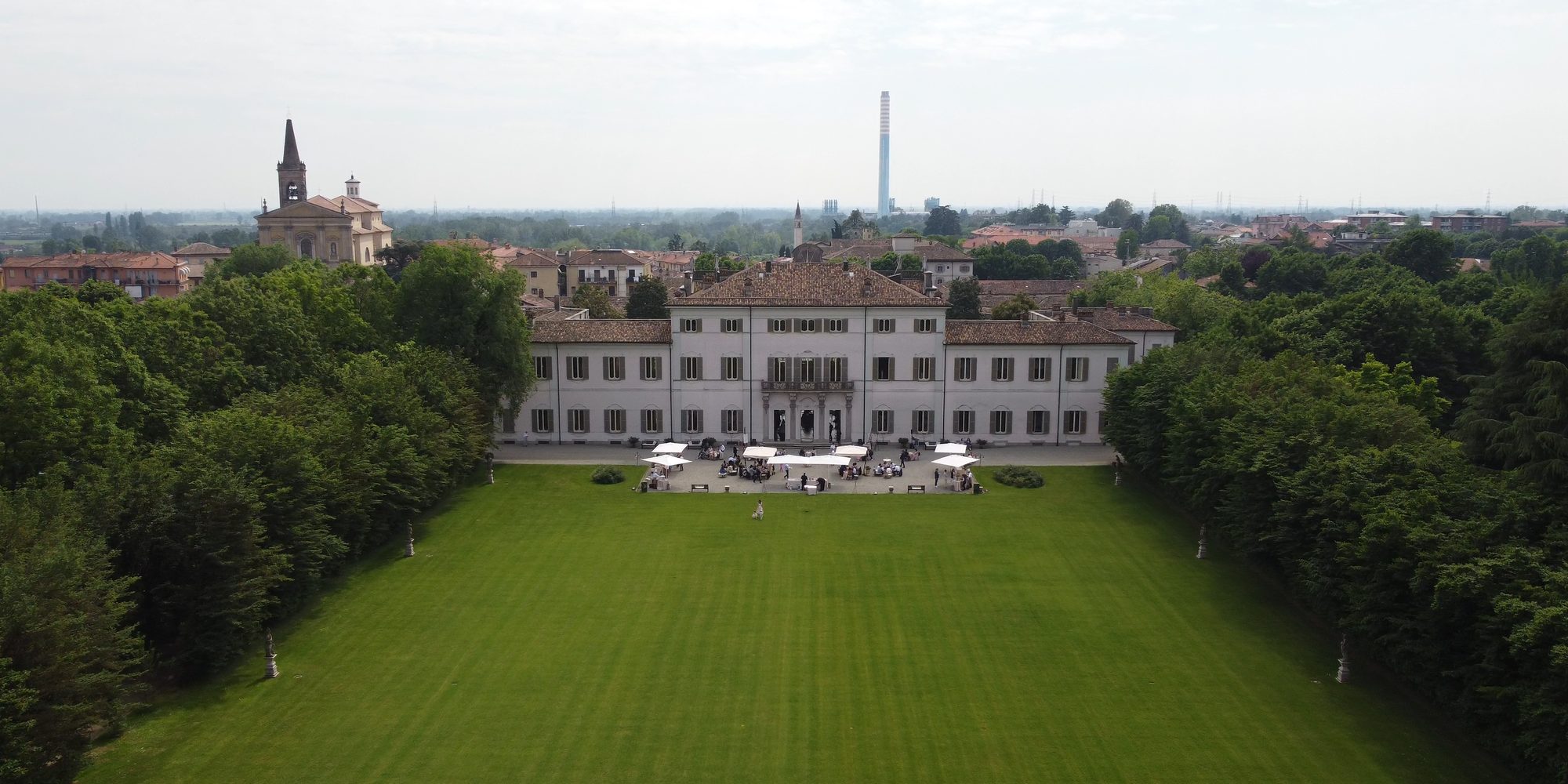 Una villa chiara in fondo a un lungo prato rasato fra due file di alberi, vista dal drone; davanti alla villa gli ombrelloni bianchi del ricevimento, dietro il paese con il campanile.