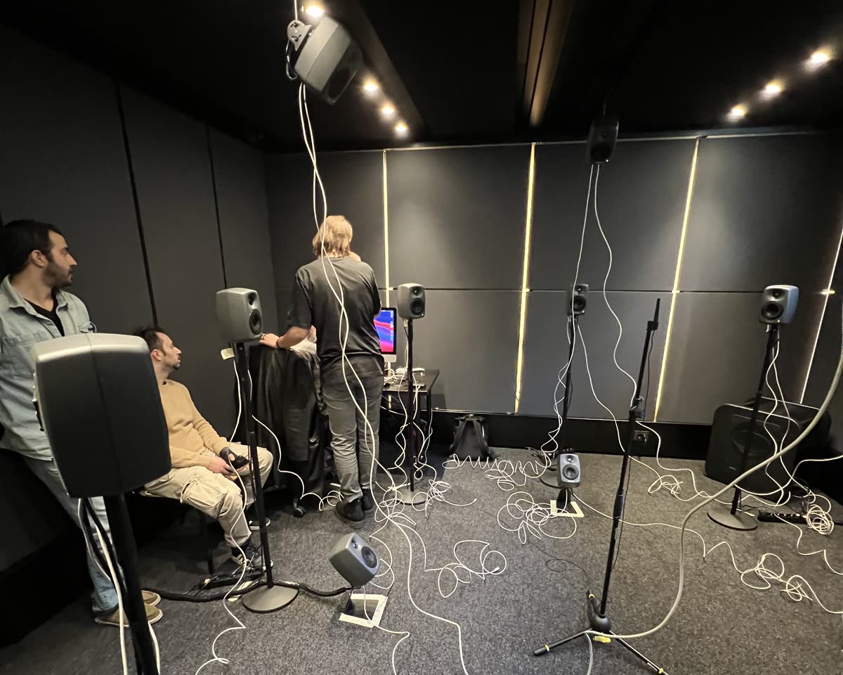 La Sala Ambisonic: pareti scure con strisce di luce, diffusori su aste e appesi al soffitto, cavi bianchi sul pavimento, tre persone intorno a una postazione.