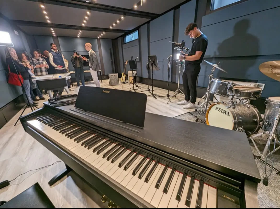 La Hall di XNL Musica: in primo piano un pianoforte digitale e una batteria, sullo sfondo un gruppo di persone, leggii, microfoni e pareti blu trattate acusticamente.