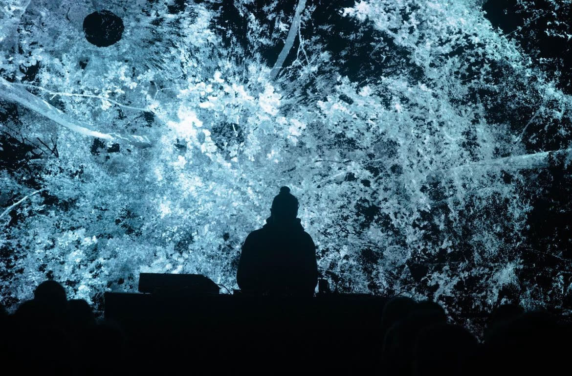 Ryoichi Kurokawa in controluce alla sua postazione, davanti a un grande schermo con una foresta ricostruita da una scansione laser, fatta di punti chiari; in basso le teste del pubblico.
