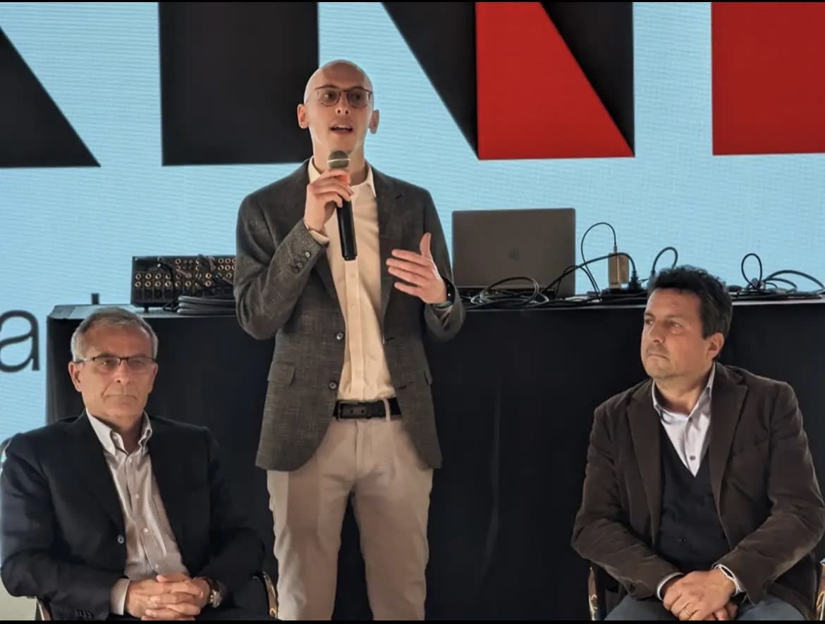 Gianmarco Romiti in piedi, con il microfono in mano, presenta gli spazi; ai lati due relatori seduti, dietro di lui il marchio di XNL.