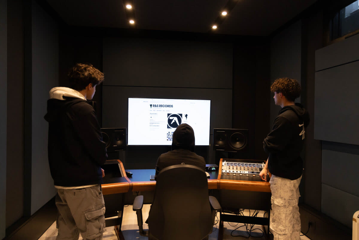 La regia di XNL Musica: la scrivania in legno con il mixer, due monitor da studio ai lati dello schermo, le pareti scure trattate acusticamente e tre studenti al lavoro.