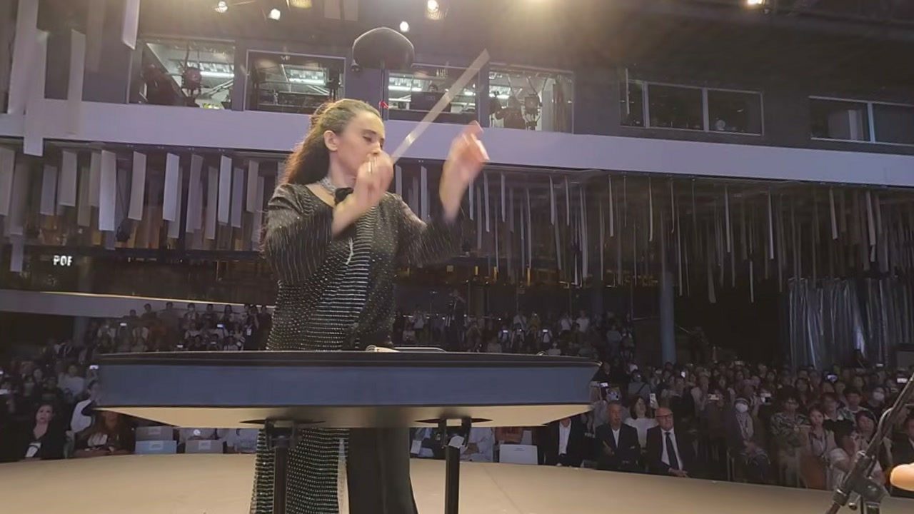 Gianna Fratta conducts from the podium, baton raised; behind her, the audience of the National Day Hall.