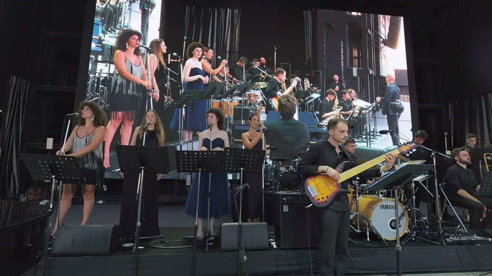 The Orchestra Nazionale Jazz dei Conservatori Italiani on the Arena Matsuri stage: the singers in a row at their music stands, the bass player, drums and horns, and behind them the big screen filming them.