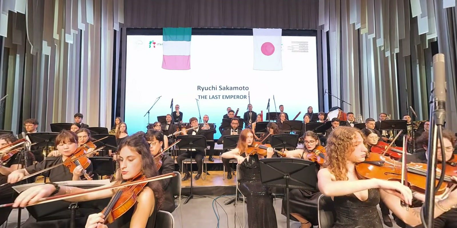 Dentro l’Orchestra Sinfonica Nazionale dei Conservatori, nella National Day Hall di Expo 2025 Osaka: violini in primo piano e, sullo schermo, le bandiere d’Italia e del Giappone con «Ryuichi Sakamoto, The Last Emperor». Un fotogramma della nostra ripresa a 360°.