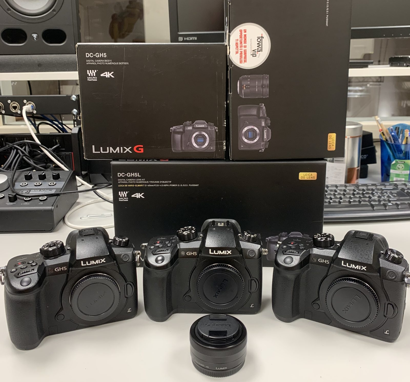 Tre Lumix GH5 in fila su un tavolo bianco, con un’ottica davanti. Dietro, le scatole: una DC-GH5 solo corpo e due DC-GH5L con lo zoom Leica 12-60 mm.