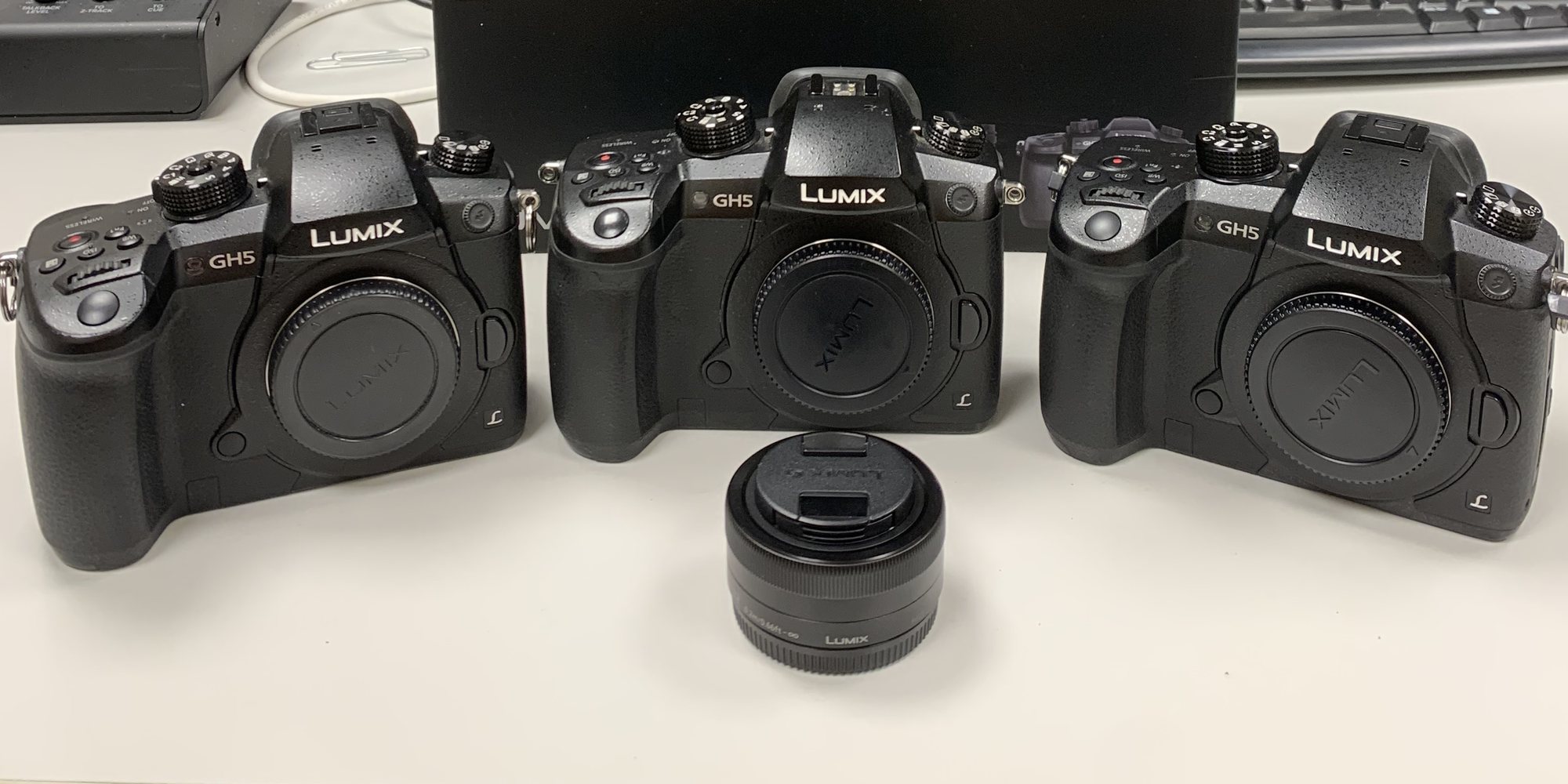 Tre Panasonic Lumix GH5 in fila su un tavolo, con un’ottica davanti; dietro, la scatola di una DC-GH5L.