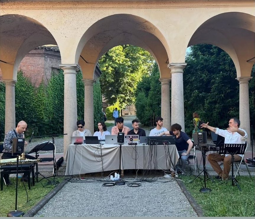 Under the portico of the Galleria Ricci Oddi cloister, a table with the computers and instruments of the live set, the musicians seated and the audience around them.