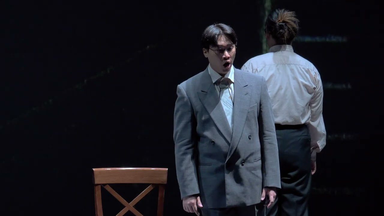 A singer in a grey suit on the dark stage of the Teatro Municipale, in front of the video set design.