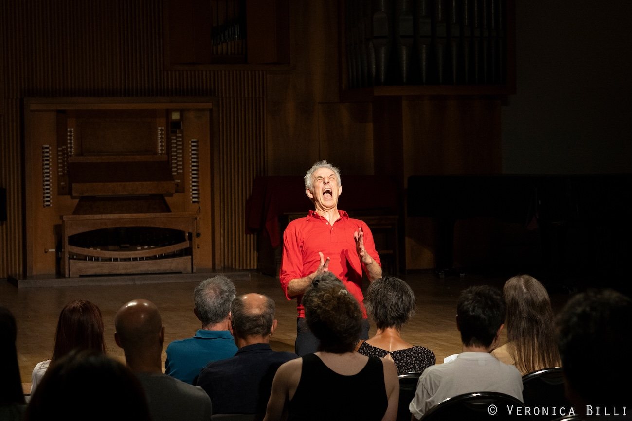 Xavier Le Roy, in a red shirt, conducts the audience seated in the hall of the Nicolini Conservatory; behind him, the organ.