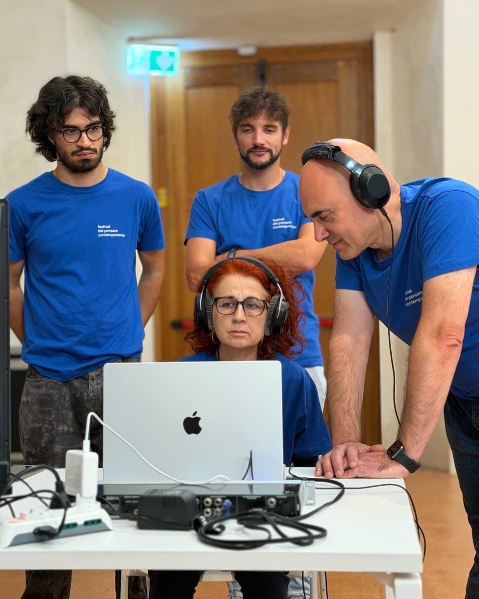 Una donna con le cuffie siede davanti a un portatile collegato a un’interfaccia audio; intorno tre persone con la maglietta blu del festival, una delle quali ascolta con un’altra cuffia.